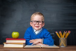 Happy cute clever boy is sitting at a desk in a glasses and smiling. Child is ready to answer with a blackboard on a background. Ready for school. Back to school. Apple and books on desk