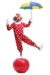 Full length portrait of a funny clown holding an umbrella and standing on a giant ball isolated on white background
