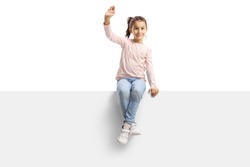 Happy little girl sitting on a panel and waving at the camera isolated on white background