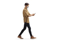 Full length profile shot of a young man with a medical face mask walking and typing on a mobile phone isolated on white background