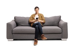 Happy young man sitting on a gray sofa and typing on a mobile phone isolated on white background