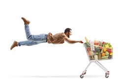 Casual man flying and holding onto a shopping cart filled with food products isolated on white background