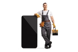 Full length portrait of a repairman in a uniform holding a tool box and leaning on a big smartphone isolated on white background