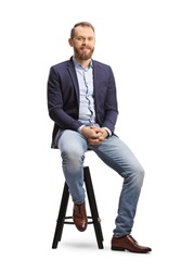 Full length portrait of a man in a suit and jeans sitting on a bar chair isolated on white background