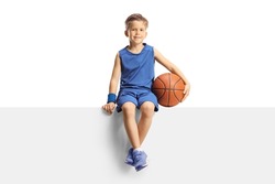 Smiling boy in a jersey holding a basketball and sitting on a blank panel isolated on white background