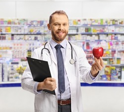 Young male pharmacist holding a clipboard and a red apple inside a pharmacy