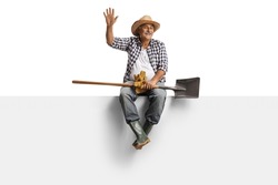 Full length portrait of a mature farmer with a spade sitting on a blank panel and waving isolated on white background