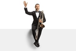 Male musician sitting on a blank panel with a trombone and waving isolated on white background