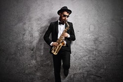 Jazz musician playing a saxophone and leaning against a rusty gray wall
