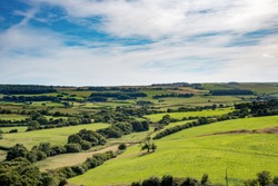 Rolling fields and trees of lush green British countryside stretch to the horizon under wispy clouds and blue sky