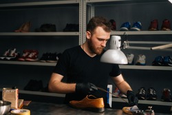 Portrait of shoemaker shoemaker wearing black gloves inserts wooden shoe pad into worn light brown leather shoes to be repaired in dark craft shoe shop. Concept of cobbler artisan work.
