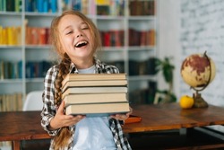 Medium shot portrait of laughing elementary child school girl holding stack of books in library at school, looking at camera. Cute primary pupil schoolgirl standing on blurry background of bookshelf.