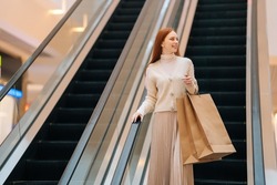 Low-angle view of smiling young woman holding on escalator handrail and riding escalator going up in shopping mall, looking away, paper bags with purchases in hands, blurred background.