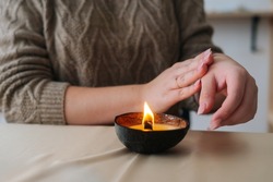 Close-up cropped shot of unrecognizable woman applying melted wax from burning handmade candle on hand. Closeup of female relaxing with aroma candle at home. Concept of wellness and relaxation.