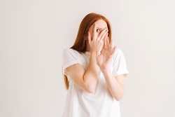 Studio portrait of frightened redhead young woman looking shy or terrified covering hiding face with hands peeping through fingers, shocked by horror movie or frightened on white isolated background.