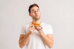 Portrait of handome young man with enjoying eating delicious slice of pizza, with closed eyes from pleasure on white isolated background. Studio shot of hungry male student eating tasty food.