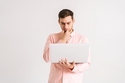 Portrait of thinking young man standing with laptop loiking on screen with confused puzzled face on white isolated background. Studio shot of pensive male posing with pc looking at screen