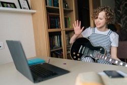 Positive preteen girl greeting and learning to play the guitar in virtual meeting together with teacher in video call. Talented kid playing guitar and watching online lessons on laptop at home.