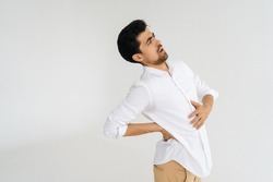 Studio portrait of unhealthy young man in shirt touching injured sore back, suffering lower lumbar discomfort, muscle pain of overwork, pinched nerve and grimacing on white isolated background.