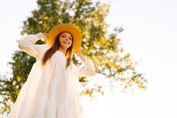 Low-angle portrait of smiling young woman in straw hat and white dress standing posing on beautiful meadow of green grass and tree looking at camera, on background of golden sunlight at summer evening
