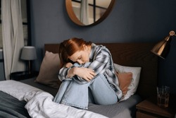 Portrait of sad upset millennial redhead female sitting on bed embrace knees lost in bad pessimistic thoughts suffer alone. Stressed young woman feel lonely desperate has broken heart.