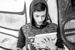 One young man, boy holding looking at tablet pad computer present touch screen isolated graffiti wall background. Negative face expressions, emotions. Technology addiction concept. Monochromatic image