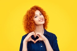 Love. Closeup portrait smiling happy young redhead curly hair woman making heart sign, symbol with hands isolated yellow wall background. Positive human emotion expression feeling life body language