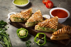 Vegetable Toste sandwich is a type of veg sandwich consisting of a vegetable filling between bread. photo is composited on grey background and wood platter with onion, capsicums  tomato, green sauces.