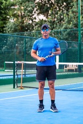 Portrait of middle aged positive male tennis player coach with racket standing at hard court.