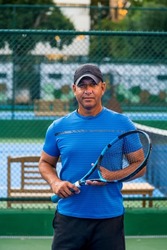 Portrait of middle aged positive male tennis player coach with racket standing at hard court.