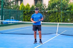Portrait of middle aged positive male tennis player coach with racket standing at hard court. sunglasses