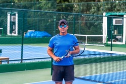 Portrait of middle aged positive male tennis player coach with racket standing at hard court. sunglasses