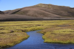 river under blue sky in the green plateau of iceland, brown hills in the background