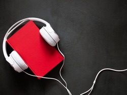 Audio book concept. White headphones and a red book on a stone black desk. Flat lay, top view, copy space