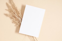 White paper empty blank, dried grass decoration on beige background. Invitation card mockup on beige table. Flat lay, top view, copy space, mockup