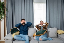 Young couple is sitting on a sofa in their apartment looking up and holding their hands to plug their ears as a neighbor upstairs is having a party and playing loud music or renovating the apartment 