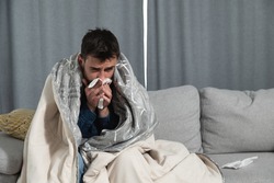 Young man suffering from a common cold and flu or allergy sit at home wrapped in blanket and wipe his nose with tissues while he have a strong headache pain, healthcare concept