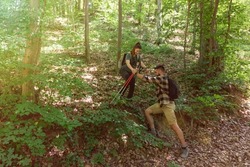 Young man helping his woman to step down on the steep hill in the forest while they hiking through nature. Couple nature lovers in the woods spending holiday like hikers. With film grain