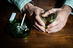 Hands of alcoholic man holding a glass with alcohol drink with smoking cigarette in the ashtray for addiction concept. Male addict drinking alcohol and smoke cigarette