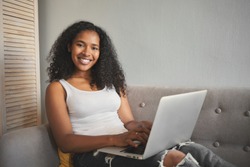 Modern technology, communication and electronic gadgets concept. Picture of beautiful positive young dark skinned woman copywriter working distantly, sitting on couch with portable computer on her lap