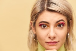 Close up highly detailed image of beautiful blonde young woman wearing bob hairstyle, nose ring, round earrings and pink eye-shadows looking at camera, having serious thoughtful look, touching chin