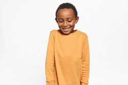 Adorable Afro American kid in yellow sweater looking down shyly having timid facial expression, smiling broadly posing against studio background with copy space for your advertising content