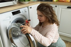 Concentrated young woman with screwdriver repairing broken washing machine. Young female fixing washer. Breakdown and repair of household appliances. Kitchen interior on blurred background
