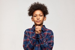 Handsome sincere African boy of 12-years-old with afro haircut standing against studio wall with fists clenched in pray, looking at camera with calm face expression. Body language, gestures and signs