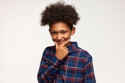 Crafty tricky boy in red and blue flannel shirt touching his chin planning joke at his friends or funny surprise, having dark skin and afro hairstyle, standing against white studio wall