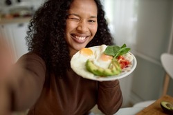 Cute young lovely adorable female of black ethnicity doing selfie over kitchen background holding plate with colorful healthy and nutritious breakfast of eggs and vegetables, winking at camera