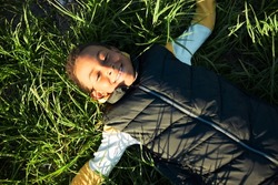Upper view of happy carefree black boy kid having rest after active outdoor games lying on green juicy grass in city park, enjoying warm water, blue sky and tranquility of moment. Happy childhood