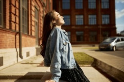 Side view outdoor image of gorgeous student girl with closed eyes in denim outfit leaning on edging at city streets taking sun bathing with face turned to sunlight, enjoying warm day and freedom