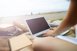 Outdoor summer shot of college student girl keyboarding on laptop pc, working on course paper, sitting alone on the riverbank with many books and copybook. People, education and technology concept
