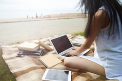 Cropped view of unrecognizable young brunette female student studying by the river, sitting on cloth with textbooks, using tablet and laptop with blank copy space screens for your information